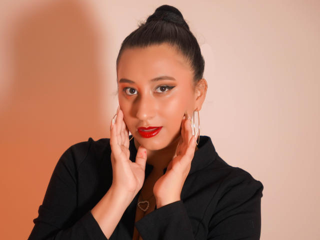 Catalina Rivero, met haar handen voor haar gezicht, een zwarte knot, rode lipstick, oorringen, een zwarte top, een hartvormige ketting, en een sexy, zelfverzekerde glimlach. Haar borsten zijn klein, haar vagina is niet zichtbaar..