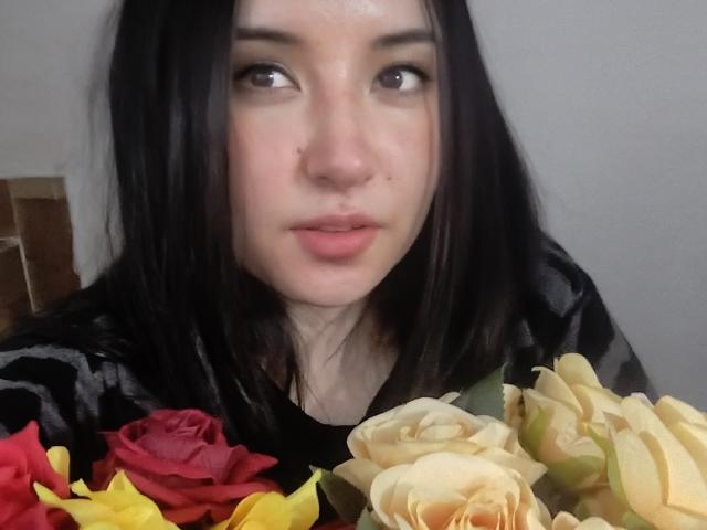 SusanMonroy with black hair, pale skin, and pouty lips, holding a bouquet of red, yellow, and beige roses. She's wearing a black and gray striped shirt.