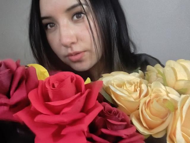 SusanMonroy with black hair, looking at camera, surrounded by red and yellow roses. Her face is slightly blurred, focusing on her expression and the flowers in the foreground.