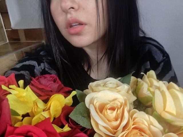 SusanMonroy with black hair, pink lips, and a black patterned top, holding a bouquet of yellow, red, and cream roses, showing her face and part of her chest.