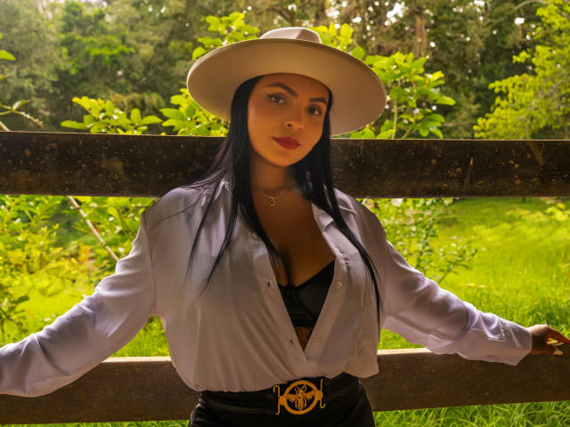 JessicaFiore with long black hair, wearing a white hat, open shirt revealing black bra, standing against a wooden fence, arms out, sunlit green background, looking at camera.