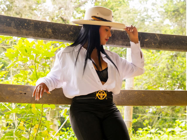 Jessica Fiore, con cabello negro, camisa blanca, un sujetador de encaje negro, pantalones negros y un cinturón dorado, lleva un sombrero blanco, apoyada en una valla de madera, con la luz del sol en su rostro..