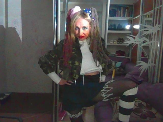 LilaniLoverX with long braids, red lipstick, camo jacket, white shirt, black skirt, and striped thigh-highs, standing with one leg up, in a room with a mirror and bookshelf.