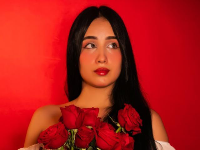 Valentina Risso, com cabelos longos e pretos, batom vermelho, segurando rosas vermelhas contra um fundo vermelho, olhando ligeiramente para o lado, olhos vermelhos, blusa branca com um ombro descoberto..