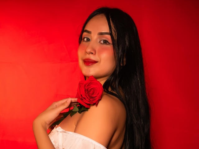 Valentina Risso, met lang zwart haar, rode lippenstift, die een rode roos tegen haar schouder houdt, gekleed in een witte top met open schouder, lachend naar de camera, rode achtergrond..