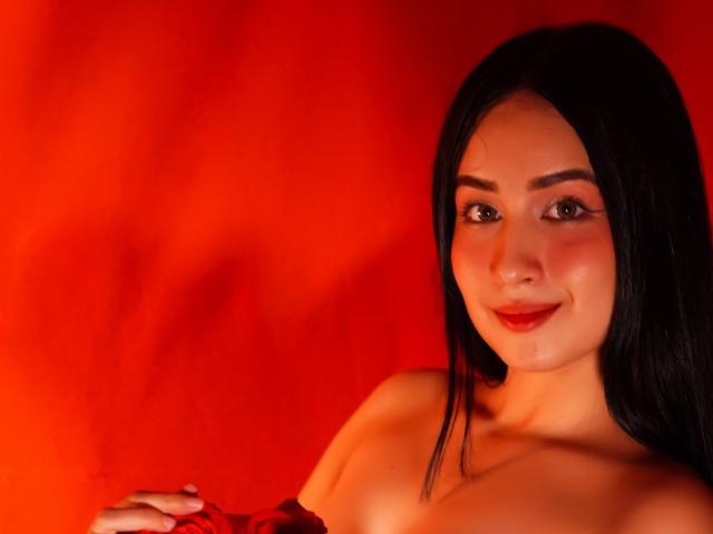 ValentinaRisso with long black hair, smiling, red lipstick, and bare shoulders, against a red background, showing off her tits and a cute, sexy face.