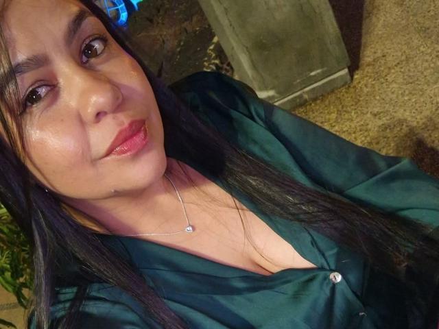 XBigTitxx has medium tits, a cute face, and long black hair. She's wearing a green satin shirt, a silver necklace, and shiny pink lipstick, smiling at the camera.
