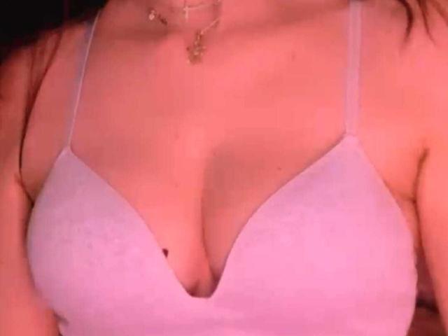 BlueNightX's pink bra-covered tits, showing a deep cleavage with a small black nipple visible. She's wearing a delicate necklace. The image has a pinkish-red filter.