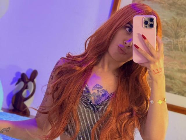 LunaDesire with long red hair, wearing a sparkly top, holding a pink phone, red nails, tattoos, and a bracelet, taking a mirror selfie. Boobs visible, purple light.