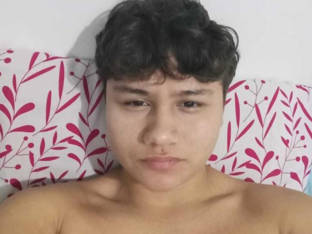 HottieFuck with short curly black hair, brown eyes, and a neutral expression, shirtless, lying on a pillow with pink floral patterns. Skin is light brown. Face is centered.