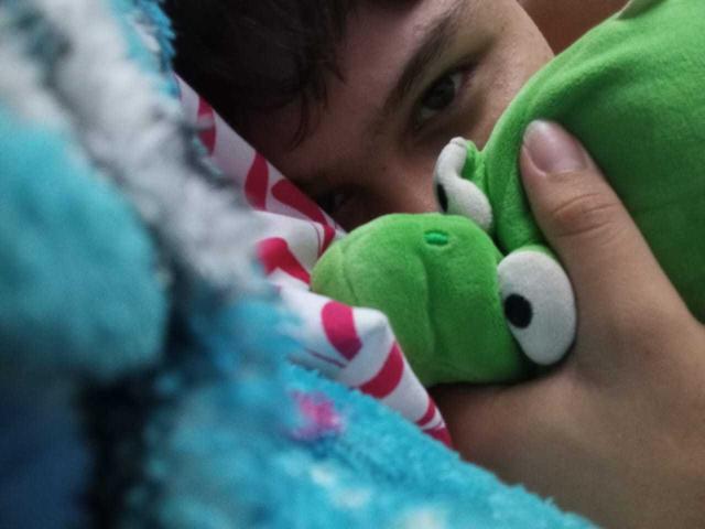 HottieFuck's face, eyes half-open, holding a green stuffed toy with white eyes. Blue and white striped blanket in foreground, blue fuzzy blanket partially visible. Close-up shot.