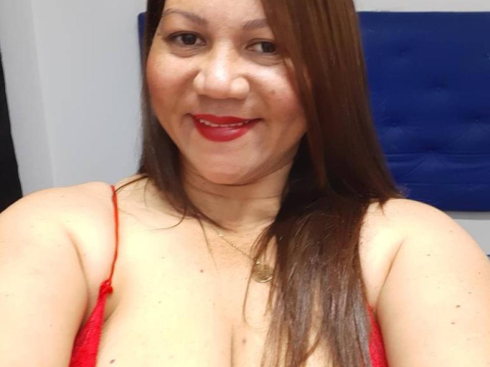 KattyLatinn with red lipstick, smiling, brown hair, wearing a red spaghetti strap top, showing her big boobs, light skin, and a gold necklace.
