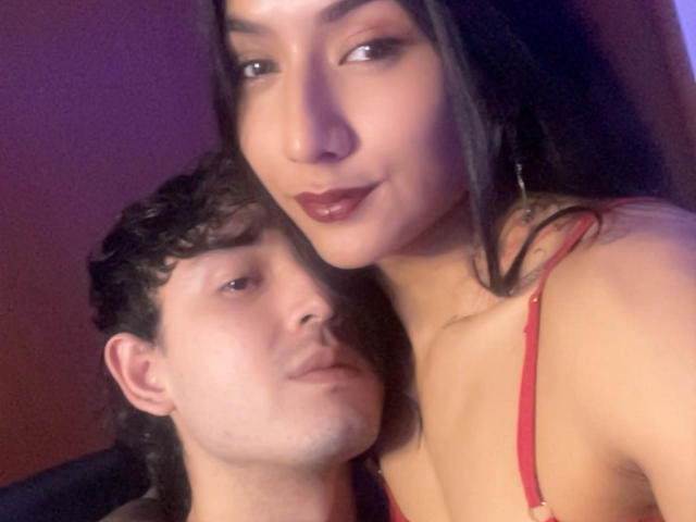 A close-up selfie of two people. TheJoestars, a woman with long dark hair, olive skin, and red lipstick, leans over a man with curly dark hair and fair skin. She wears a red bra, and her lips are slightly parted. The man looks up at her with a neutral expression. The woman's face is in the foreground, while the man's is partially obscured. The lighting is soft, creating a subtle purple hue. TheJoestars' makeup is subtle yet effective.