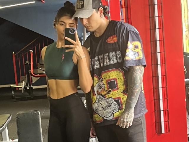 TheJoestars with medium tits and toned abs in a green sports bra, black leggings, standing next to TheJoestars in a grey shirt with "Bears" and tattoos, holding a phone.