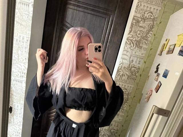 SlankyMic-hot with pink hair, black top, and black loose shirt, holding phone, taking mirror selfie, showing tits, in dark room, with fridge and patterned wall.