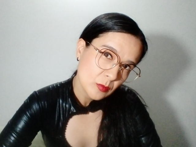 AileenRamirezW with black leather outfit, red lipstick, and glasses, has medium tits, dark hair, and a serious look, against a gray background.