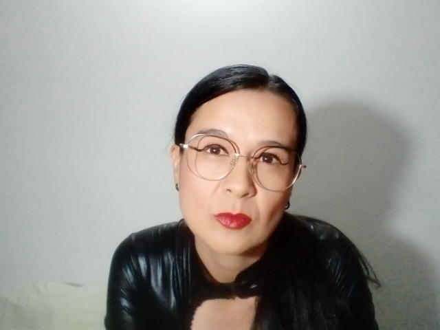 AileenRamirezW with black hair, red lipstick, and glasses, wearing a black leather jacket, looking directly at the camera with a serious expression.