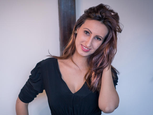 LysAddiction has brown hair, a black dress with a V-neck, and is smiling with her hand in her hair. Her tits are visible and she has pink lipstick.