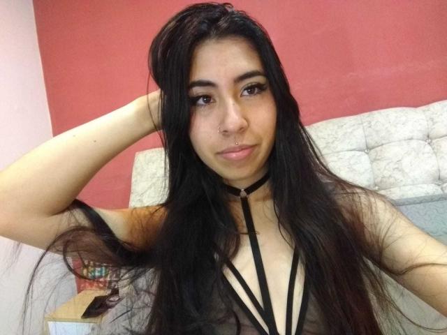 LinaSpearce with long black hair, nose ring, and a black strappy top, raising her arm, looking at the camera, showing her tits. She has a slight smile.