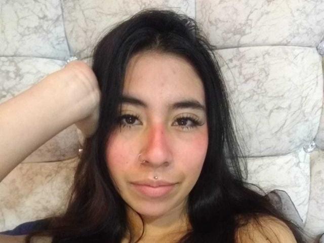 LinaSpearce mit dunklem Haar, Nasenring und Septum-Piercing, lächelnd, Hand im Haar, liegend auf einem Kopfbrett mit Marmormuster, blickt in die Kamera..
