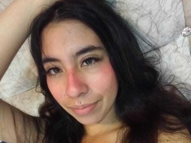 LinaSpearce has dark, wavy hair, brown eyes, a small nose ring, and a piercing on her lower lip, lying with arms above her head, smiling slightly.