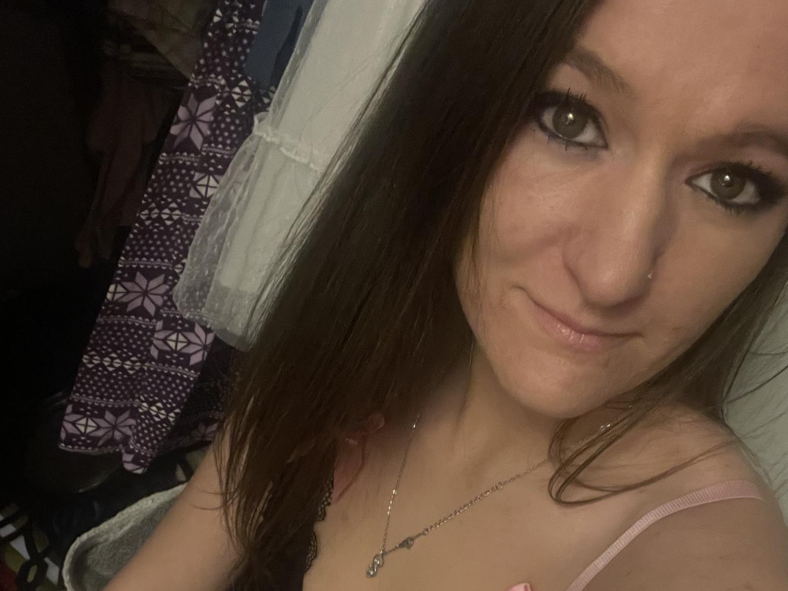 MaddyLoue mit langen braunen Haaren, Nasenpiercing und einem leichten Lächeln, trägt ein pinkes Oberteil, hat kleine Brüste und eine silberne Halskette und blickt mit braunen Augen in die Kamera..