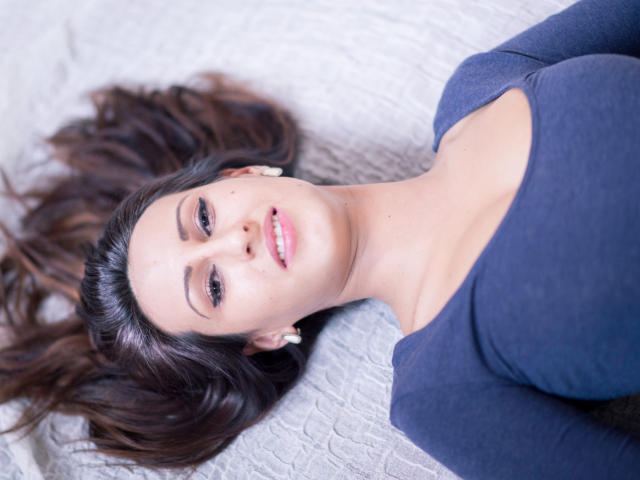 LysAddiction lying on her back, dark brown hair spread out, wearing a blue top with a low neckline, showing her tits, pink lips, earrings, and a slight smile.