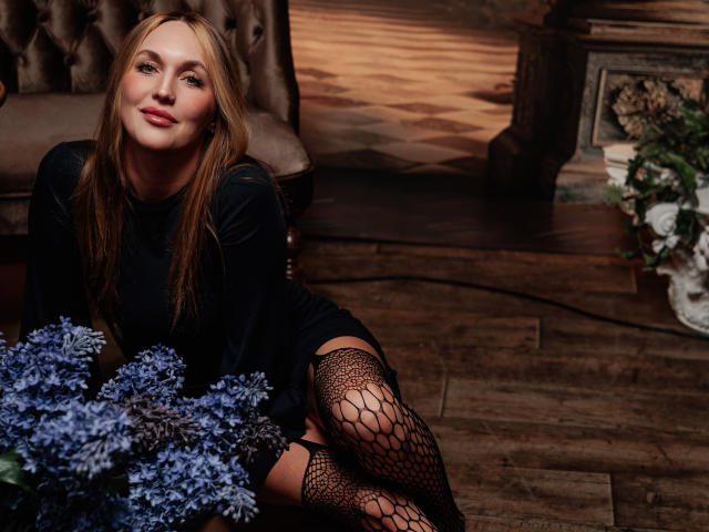 EvelynCute with long blonde hair, wearing a black long-sleeve top and fishnet stockings, sits on a wooden floor. She leans slightly forward with a relaxed smile. Her right leg is crossed over her left. Blue flowers are in the foreground. The background has a tufted chair, a wooden floor, and a stone fireplace with flowers. Warm lighting creates a cozy atmosphere. The image has a realistic, detailed style with soft shadows and highlights.