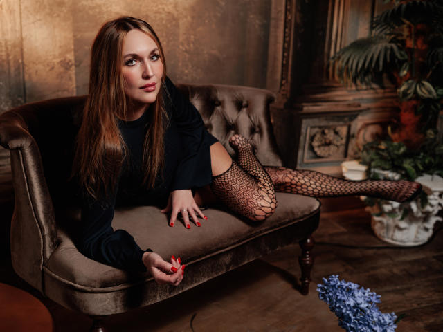 EvelynCute is a young woman with long brown hair lying on a brown velvet couch. She wears a black long-sleeve top and fishnet stockings with a floral pattern. Her red-painted nails rest on the couch. The room has a rustic, ornate wooden wall and a potted plant with dark green leaves. A blue floral arrangement is on the floor to the right. The lighting is warm, highlighting EvelynCute's fair skin and relaxed expression.