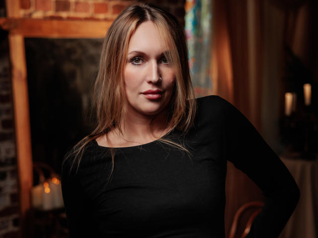 EvelynCute has light brown hair, straight and slightly parted in the middle. She wears a tight black long-sleeve top, highlighting her medium-sized breasts. Her blue eyes are slightly downcast, and her lips are subtly pink. The lighting is warm, casting soft shadows on her face. The background includes a brick wall and blurred candles, creating a cozy, intimate atmosphere. The image has a slightly overexposed effect, emphasizing EvelynCute's facial features. The composition focuses on her upper body and face.