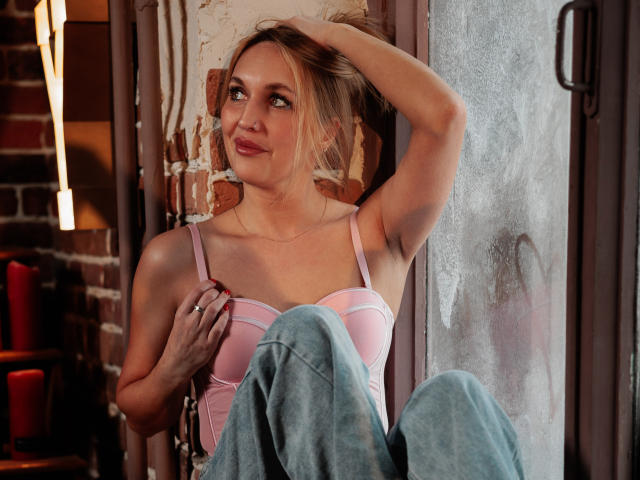 EvelynCute with blonde hair, pink bra, and blue jeans, sits against a wall, hand in hair, smiling, looking to the side, showing small tits, relaxed, and cute.