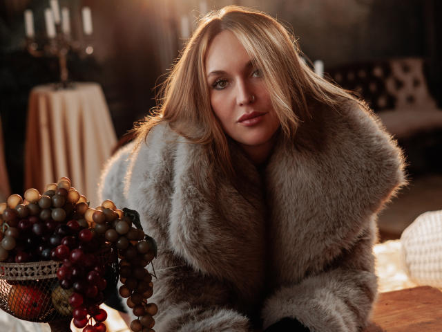 EvelynCute with blonde hair, green eyes, and a sexy look, wearing a fluffy grey fur coat, leaning forward, showing off her tits, with a grape bowl in the foreground.