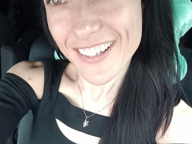 BellaJulls smiling with white teeth, black hair, and a cute nose, wearing a black top and seatbelt, necklace with a small "N" pendant, in a car.