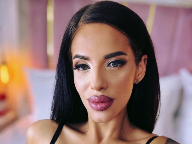 LuxuryAnne has smooth, tan skin, dark straight hair, and full, pink lips. She's wearing dark eyeliner and a black top, with small earrings. Her ass is not visible.