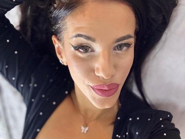 LuxuryAnne with dark hair, wearing a black polka dot shirt, has pink lipstick, eyeliner, and a butterfly necklace, lying down with arms above her head.