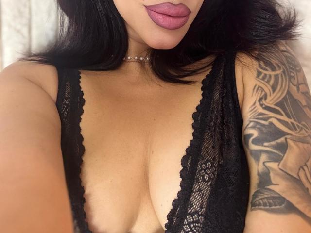 LuxuryAnne with dark hair, purple lipstick, black lace top, showing her tits, tattooed arm, and a delicate choker, taking a selfie.
