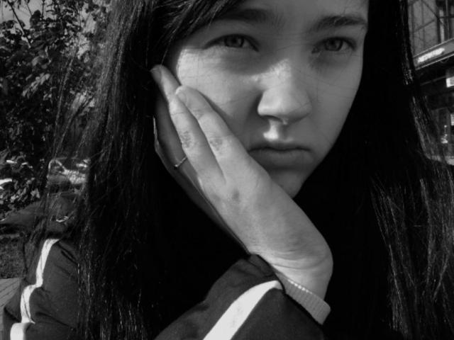 Black-and-white photo of KateSii with long dark hair, hand on cheek, nose ring, serious expression, wearing a striped jacket, fingers showing a ring.