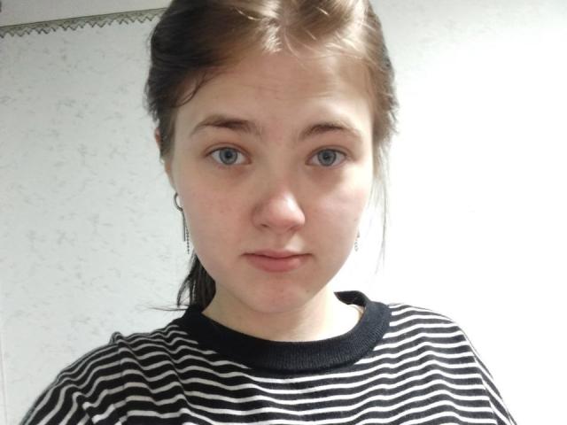 KateSii has blue eyes, light skin, and brown hair in a ponytail. She's wearing a black and white striped shirt with small earrings. Her face is slightly pouty.