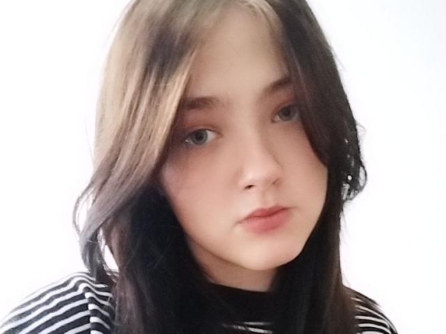 KateSii has light skin, brown eyes, and straight brown hair with blonde highlights. She's wearing a black and white striped shirt, looking serious with slightly parted lips.