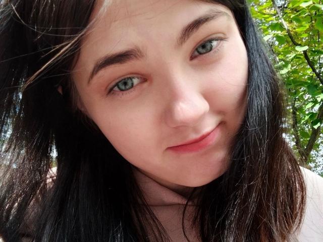 KateSii has pale skin, green eyes, and black hair, with a slight smile, looking at the camera, showing her cute face and small nose. Her tits are not visible.