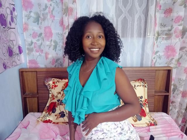 LuciaSexy69, dark-skinned woman with curly black hair, smiling, wearing a teal ruffled top and white floral skirt, standing in front of a wooden bed with floral pillows.