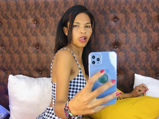 CharlineMagik with dark brown hair, tan skin, and red lipstick, taking a selfie in a checkered dress, showing her tits, with a phone in a brown, patterned headboard.