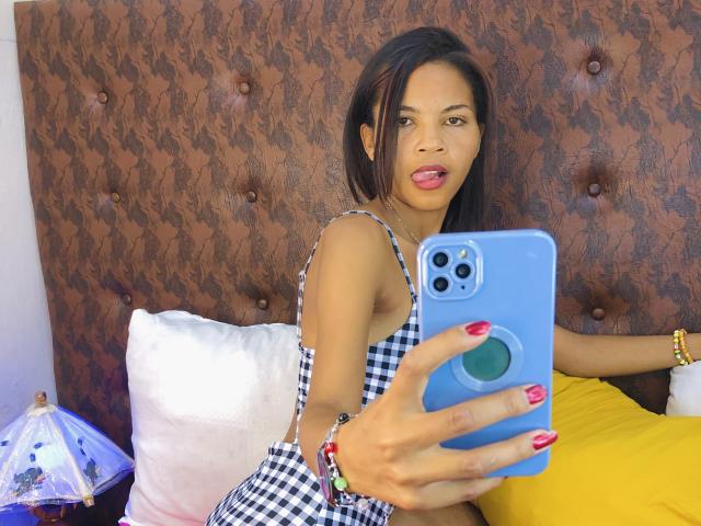 CharlineMagik taking a selfie with a blue phone, tongue out, wearing a checkered dress, red nails, and colorful bracelets, in a bedroom with a tufted brown headboard.
