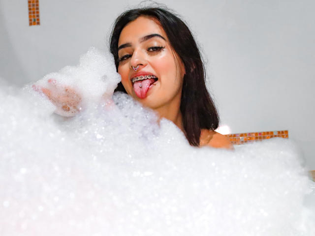 AtheneaOwenss in a bubble bath, sticking out her tongue with a nose ring, dark hair, and visible breasts covered in white foam. Her eyes are half-closed and she's smiling.