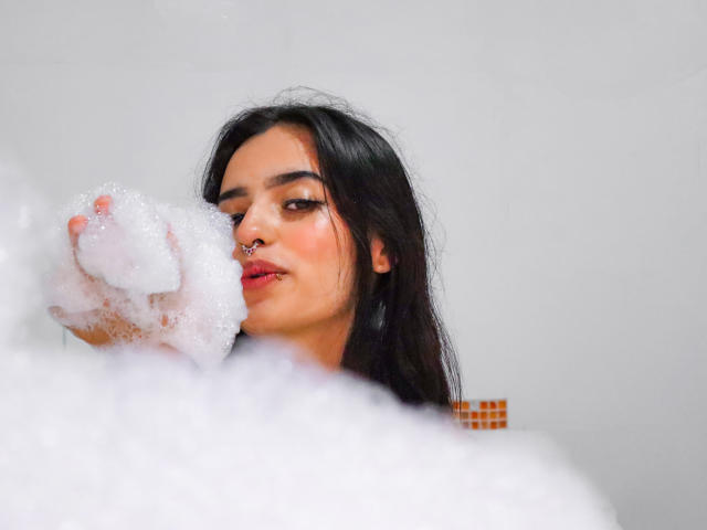 AtheneaOwenss with long black hair, nose ring, and red lipstick, holding a soapy hand in the foreground, looking slightly to the side, with a fluffy white bubble.