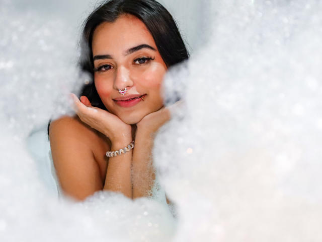 AtheneaOwenss with dark hair, nose ring, and bracelet, smiling in a bubble bath, hands under chin, looking at camera, soft white bubbles in foreground.