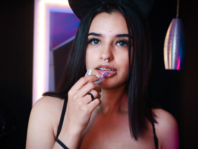 AtheneaOwenss with dark hair, blue eyes, nose ring, and white nail polish, sucking on a pink dildo, wearing a black hat and bra, in a dimly lit room.