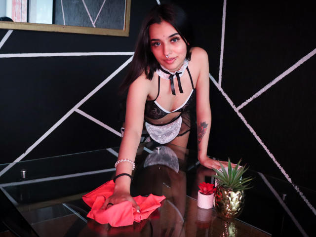 AtheneaOwenss with dark hair, medium tits, black lace lingerie, white frill, and red ribbon, cleaning glass table, red cloth, potted plant, gold vase, tattoo on right arm.