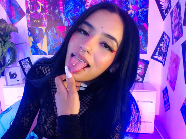 AtheneaOwenss with long black hair, pink and red eye makeup, nose and tongue piercings, sticking out tongue, holding finger to mouth, wearing black mesh top, purple and blue lighting.