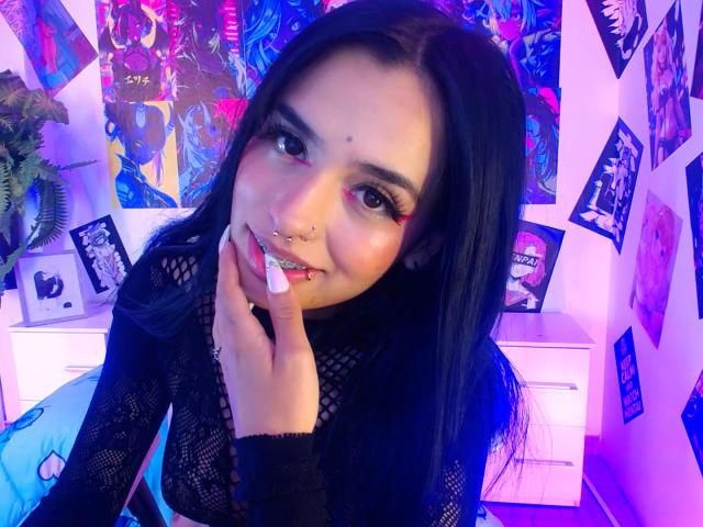AtheneaOwenss with long black hair, purple eye makeup, nose piercings, and a septum piercing, wearing a black lace top, touching her chin, in a colorful, neon-lit room.